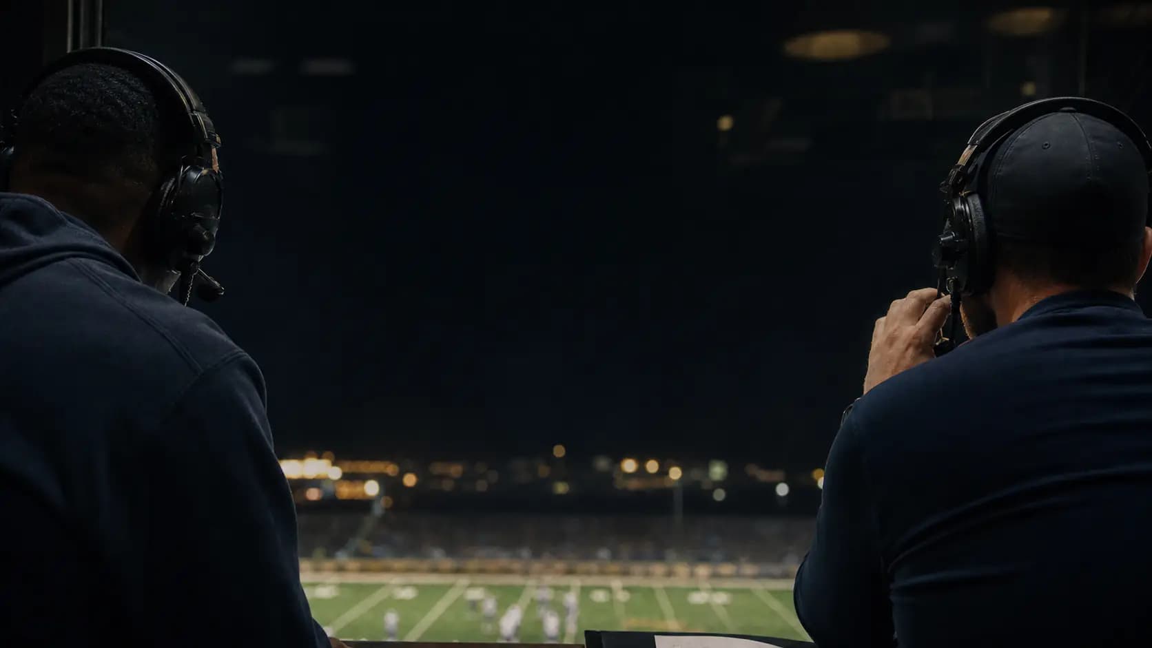 Two football analysts reviewing game film from opposite sides of a stadium booth